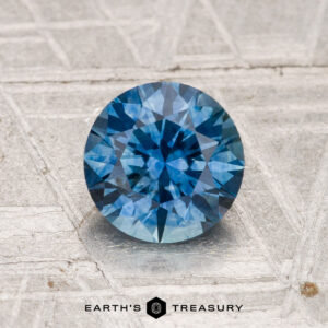 A 1.34-carat heated Montana sapphire with a brilliant round cut sits on a textured gray surface, the "Earth's Treasury" logo displayed at the bottom.