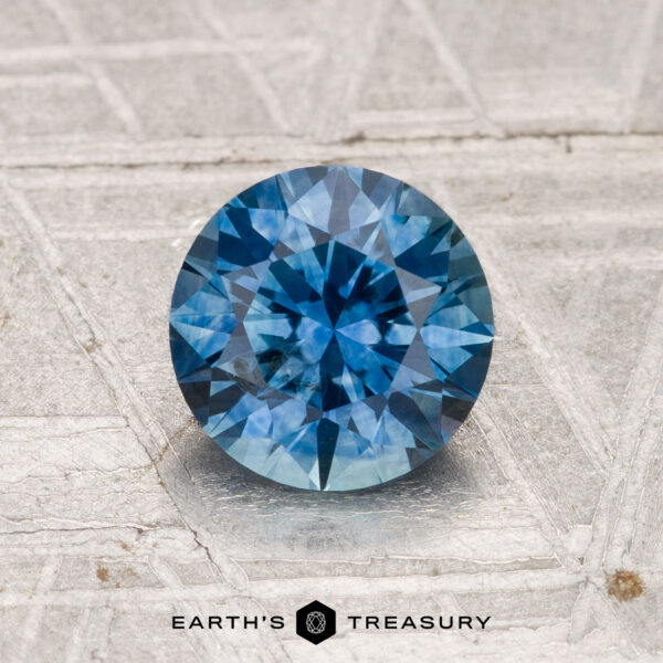1.34-Carat Montana Sapphire (Heated) A 1.34-carat heated Montana sapphire with a brilliant round cut sits on a textured gray surface, the "Earth's Treasury" logo displayed at the bottom.