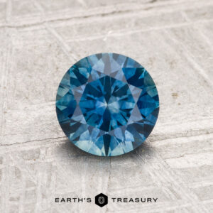 1.33-carat heated Montana sapphire in round brilliant cut shown on textured gray surface, with "EARTH'S TREASURY" text at the bottom.