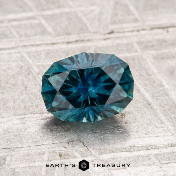 A 1.31-carat heated Montana sapphire in blue-green rests on a textured light gray surface. "EARTH'S TREASURY" appears at the bottom of the image.