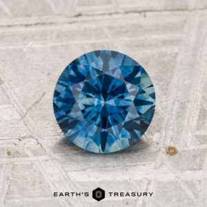 1.32-carat heated Montana sapphire in round brilliant cut displayed on a textured gray surface, with "EARTH'S TREASURY" text at the bottom of the image.