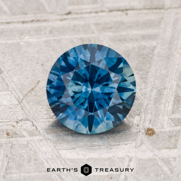 1.32-Carat Montana Sapphire (Heated) 1.32-carat heated Montana sapphire in round brilliant cut displayed on a textured gray surface, with "EARTH'S TREASURY" text at the bottom of the image.