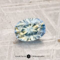 A 1.21-carat heated Montana sapphire with blue and yellow hues is displayed on a textured gray surface with the text "EARTH'S TREASURY" below.