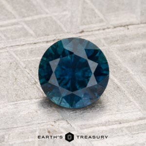 A 1.31-carat round-cut Montana sapphire (heated) is displayed on a textured metallic surface with the text "EARTH'S TREASURY" beneath it.