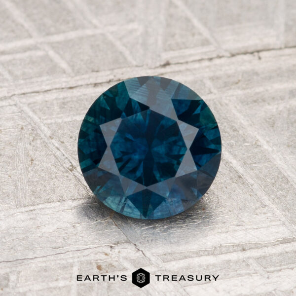 1.31-Carat Montana Sapphire (Heated) A 1.31-carat round-cut Montana sapphire (heated) is displayed on a textured metallic surface with the text "EARTH'S TREASURY" beneath it.
