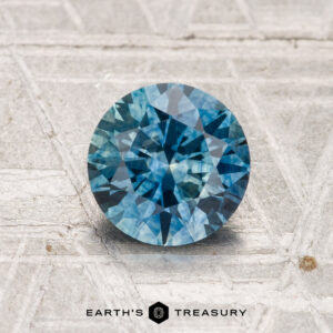 A 1.31-carat, heated Montana sapphire with a round brilliant cut is displayed on a textured, light-colored surface above the "Earth’s Treasury" logo.