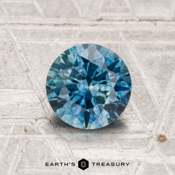 1.31-Carat Montana Sapphire (Heated) A 1.31-carat, heated Montana sapphire with a round brilliant cut is displayed on a textured, light-colored surface above the "Earth’s Treasury" logo.