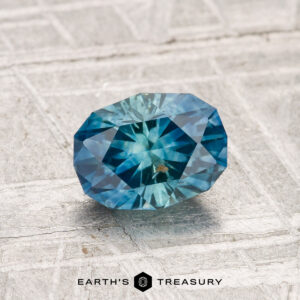 A 0.94-carat heated Montana sapphire with a faceted blue starburst pattern is displayed on a textured metallic surface, with the "Earth's Treasury" logo beneath.