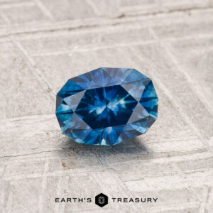 A 0.94-carat heated Montana sapphire shines on a textured gray surface, with "Earth's Treasury" displayed at the bottom.