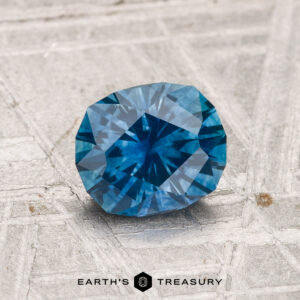 A 1.23-carat heated Montana sapphire rests on a textured, light surface with "EARTH'S TREASURY" and a hexagon logo below.