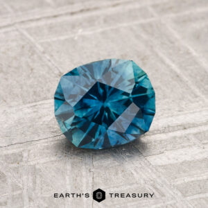 A 1.16-carat heated Montana sapphire with geometric cuts is displayed on a textured gray surface. "EARTH'S TREASURY" appears at the bottom.