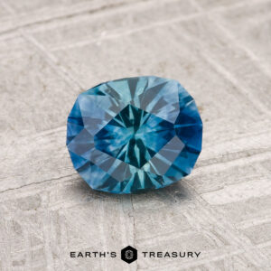 A 1.08-carat heated Montana sapphire with a square cushion cut rests on a textured gray surface. "EARTH'S TREASURY" is displayed at the bottom of the image.