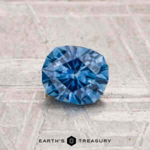A 0.81-carat heated Montana sapphire with a rich blue hue rests on a textured light gray surface, featuring the "EARTH'S TREASURY" text and a hexagon logo at the bottom.