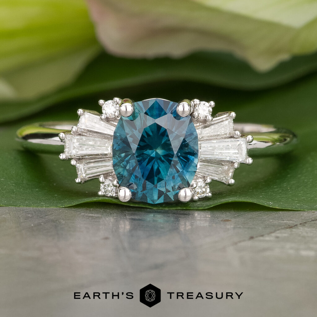 The _SNY0260-2-Edit-1200-BW ring features an oval blue gemstone at the center, accented by baguette and round clear stones, showcased against a green leaf background.