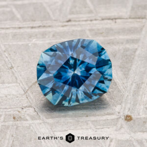 A 1.36-carat heated Montana sapphire with a starburst pattern lies on a textured gray surface, with "EARTH'S TREASURY" displayed at the bottom.