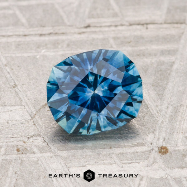 1.36-Carat Montana Sapphire (Heated) A 1.36-carat heated Montana sapphire with a starburst pattern lies on a textured gray surface, with "EARTH'S TREASURY" displayed at the bottom.