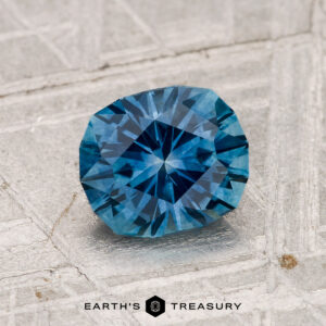 A 1.36-carat heated Montana sapphire with a faceted geometric cut rests on a textured light gray surface. The "EARTH'S TREASURY" logo is visible at the bottom of the image.