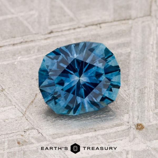 1.36-Carat Montana Sapphire (Heated) A 1.36-carat heated Montana sapphire with a faceted geometric cut rests on a textured light gray surface. The "EARTH'S TREASURY" logo is visible at the bottom of the image.