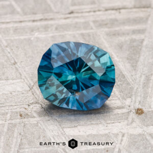 A round, faceted blue-green 1.30-carat Montana Sapphire (heated) rests on a textured gray surface with "Earth's Treasury" written below.