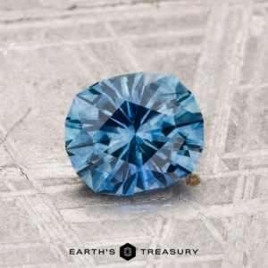 A 1.29-carat heated Montana sapphire with a faceted blue appearance is shown on a textured metallic surface, featuring "EARTH'S TREASURY" at the bottom of the image.