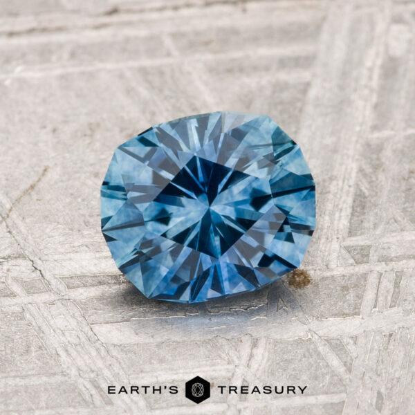 1.29-Carat Montana Sapphire (Heated) A 1.29-carat heated Montana sapphire with a faceted blue appearance is shown on a textured metallic surface, featuring "EARTH'S TREASURY" at the bottom of the image.