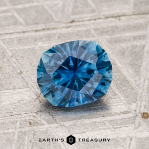 A 1.27-carat heated Montana sapphire with a blue hue rests on a textured gray surface, with the text "Earth's Treasury" at the bottom of the image.
