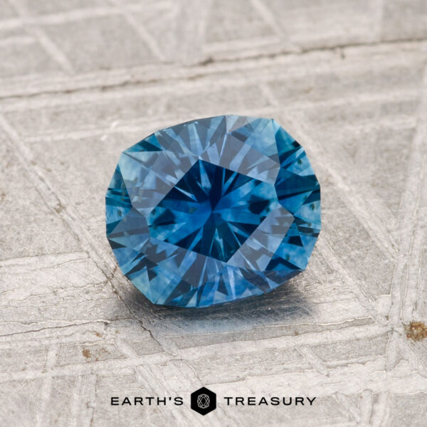 1.27-Carat Montana Sapphire (Heated) A 1.27-carat heated Montana sapphire with a blue hue rests on a textured gray surface, with the text "Earth's Treasury" at the bottom of the image.