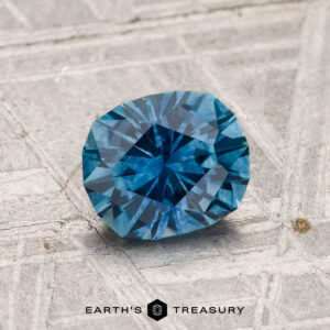 A 1.26-carat heated Montana sapphire with a blue hue rests on a textured gray surface, "EARTH'S TREASURY" inscribed below.
