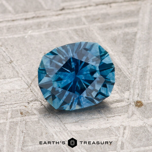 1.26-Carat Montana Sapphire (Heated) A 1.26-carat heated Montana sapphire with a blue hue rests on a textured gray surface, "EARTH'S TREASURY" inscribed below.