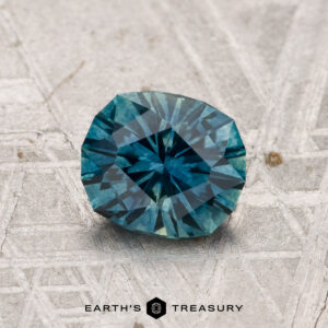 A 1.26-carat heated Montana sapphire in a round brilliant cut, showcasing a teal-blue hue, is displayed on a textured gray surface with the text "EARTH'S TREASURY" below.