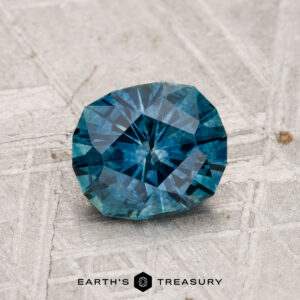 A 1.25-carat heated Montana sapphire, cut in a round geometric style, sits on a textured gray surface. The "Earth's Treasury" logo is displayed below.