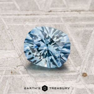A 1.24-carat heated Montana sapphire is displayed on a textured gray surface with the text "EARTH'S TREASURY" below.