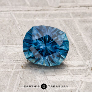 A 1.22-carat heated Montana sapphire is showcased on a textured gray surface with "EARTH'S TREASURY" at the bottom.