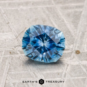 A 1.19-carat heated Montana sapphire rests on a textured grey surface, with "EARTH'S TREASURY" displayed at the bottom.