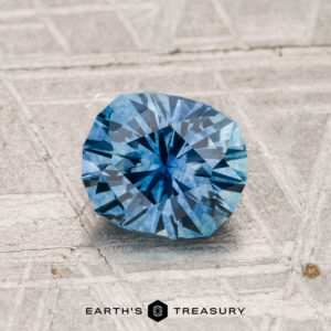 A radiant-cut, faceted blue 1.19-carat heated Montana sapphire rests on a textured gray surface, with "EARTH'S TREASURY" text displayed at the bottom of the image.