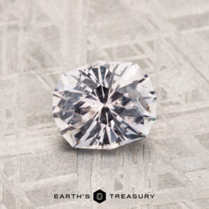 A 1.11-carat Montana Sapphire, round and faceted, rests on a textured metallic surface with "Earth's Treasury" displayed below.