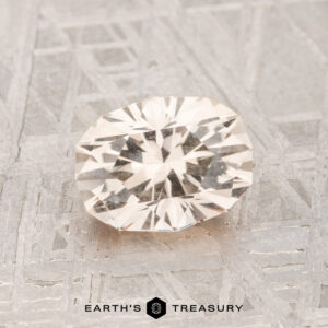 A faceted, clear 1.37-carat Montana sapphire is displayed on a textured metallic surface featuring "EARTH'S TREASURY" and a logo at the bottom center.