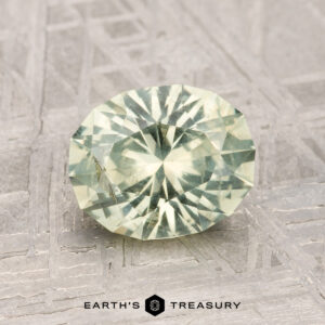 A 1.79-carat Montana Sapphire, faceted in a pale green oval, is displayed on a textured metallic surface with "EARTH'S TREASURY" shown below.