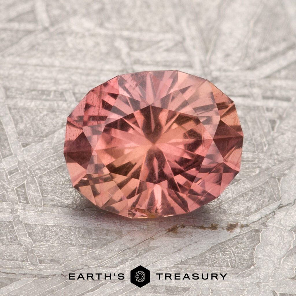 A faceted, oval-cut 0.72-Carat Padparadscha Pink Umba Sapphire rests on a light textured surface with "Earth's Treasury" below it.