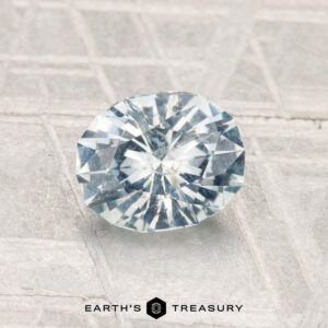 A 1.24-carat round brilliant-cut light blue Montana sapphire on a textured gray surface, featuring the "Earth's Treasury" logo and text at the bottom.