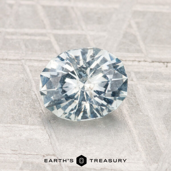 1.24-Carat Montana Sapphire A 1.24-carat round brilliant-cut light blue Montana sapphire on a textured gray surface, featuring the "Earth's Treasury" logo and text at the bottom.