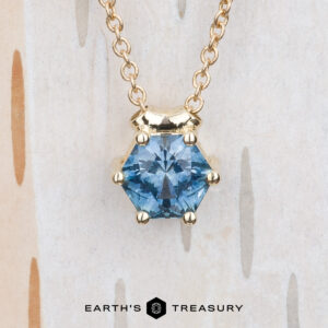The BYO "Zephyr" necklace in 14K yellow gold, featuring a 0.66-carat hexagon Montana sapphire pendant, is displayed on a light wooden background. The Earth's Treasury logo is shown at the bottom.