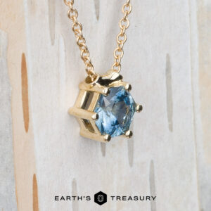 The BYO "Zephyr" necklace in 14K yellow gold, featuring a 0.66-carat hexagon Montana sapphire pendant, is displayed on a light wooden background. The Earth's Treasury logo is shown at the bottom.