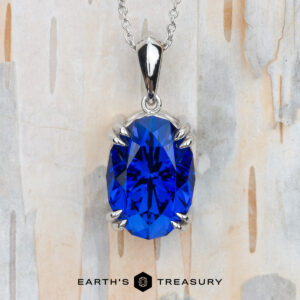The "Aria" pendant in platinum with an 8.05-carat Royal Blue Tanzanite is shown on a light wood background, with the text "EARTH'S TREASURY" displayed below.