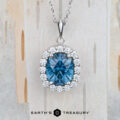 Custom Halo Pendant in 14K white gold featuring a 2.59-carat oval Montana sapphire, accented by a halo of clear stones on a silver chain. Displayed on a light textured background with the "Earth's Treasury" logo at the bottom.