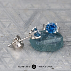 A pair of EA-MS-403 4.0mm silver stud earrings with round blue gemstones, one upright and one flat, displayed on a textured surface with a blue stone.