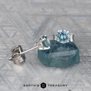 A pair of EA-MS-401 4.0mm silver stud earrings with round blue gemstones, one on a rough blue stone, displayed on a textured gray surface. Earth’s Treasury logo appears at the bottom.