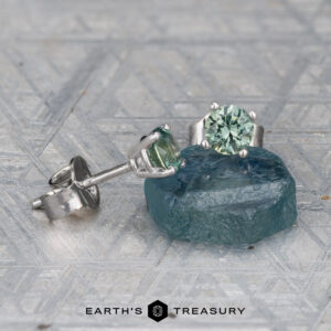 EA-MS-405 4.0mm green gemstone stud earrings in silver settings, shown with a rough blue gemstone on a textured surface. "Earth's Treasury" logo appears at the bottom.