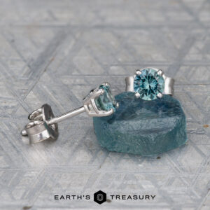 A pair of EA-MS-404 4.0mm blue gemstone stud earrings in silver settings sits beside a rough blue stone on a textured surface, with the "Earth's Treasury" logo displayed below.