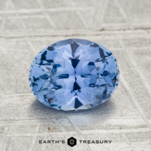 A faceted round 2.06-carat heated Ceylon sapphire is displayed on a textured surface with the text "EARTH'S TREASURY" beneath it.